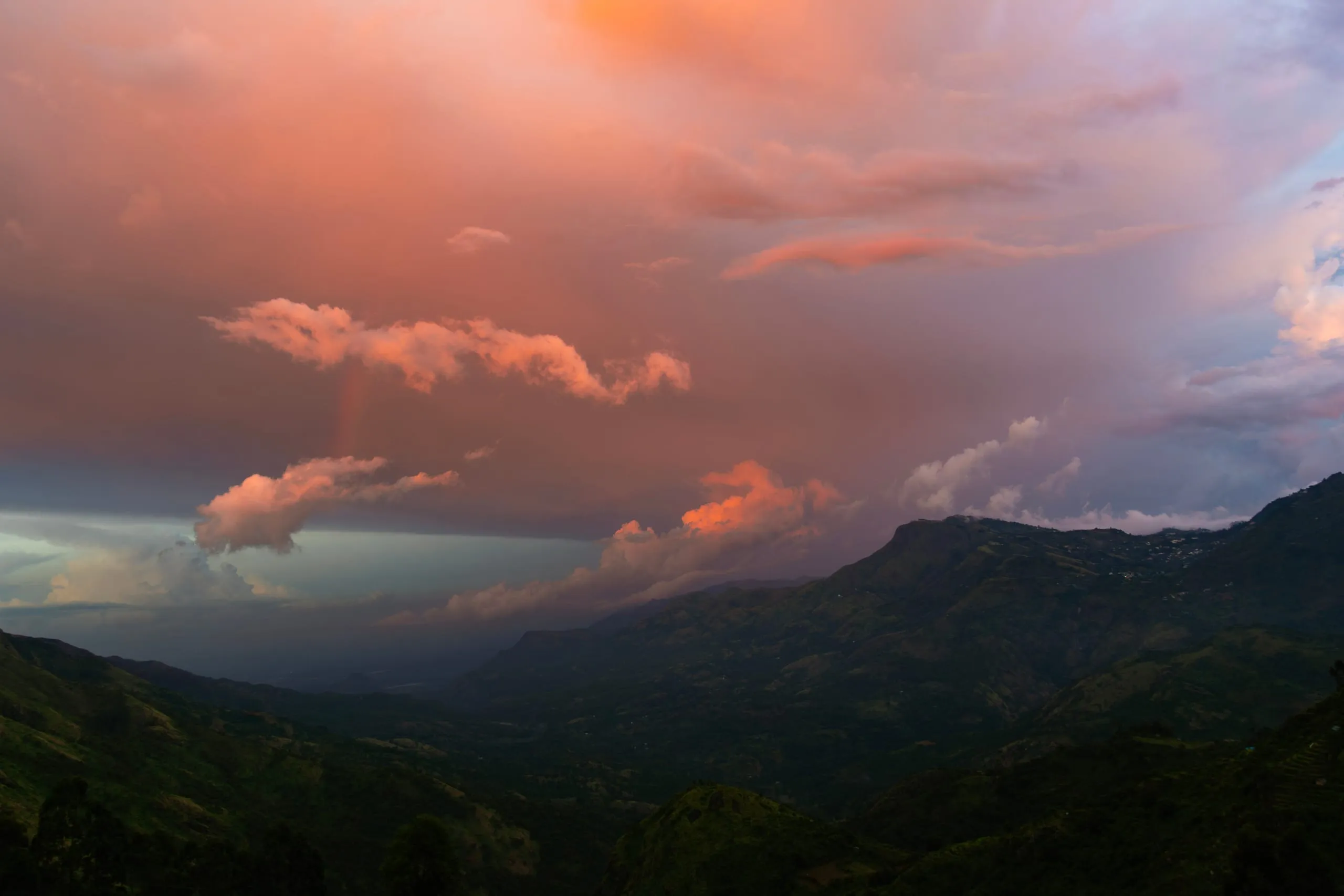 Storm cloud lit orange and pink at sunset above a dark green valley, with rain falling in a shaft on the left